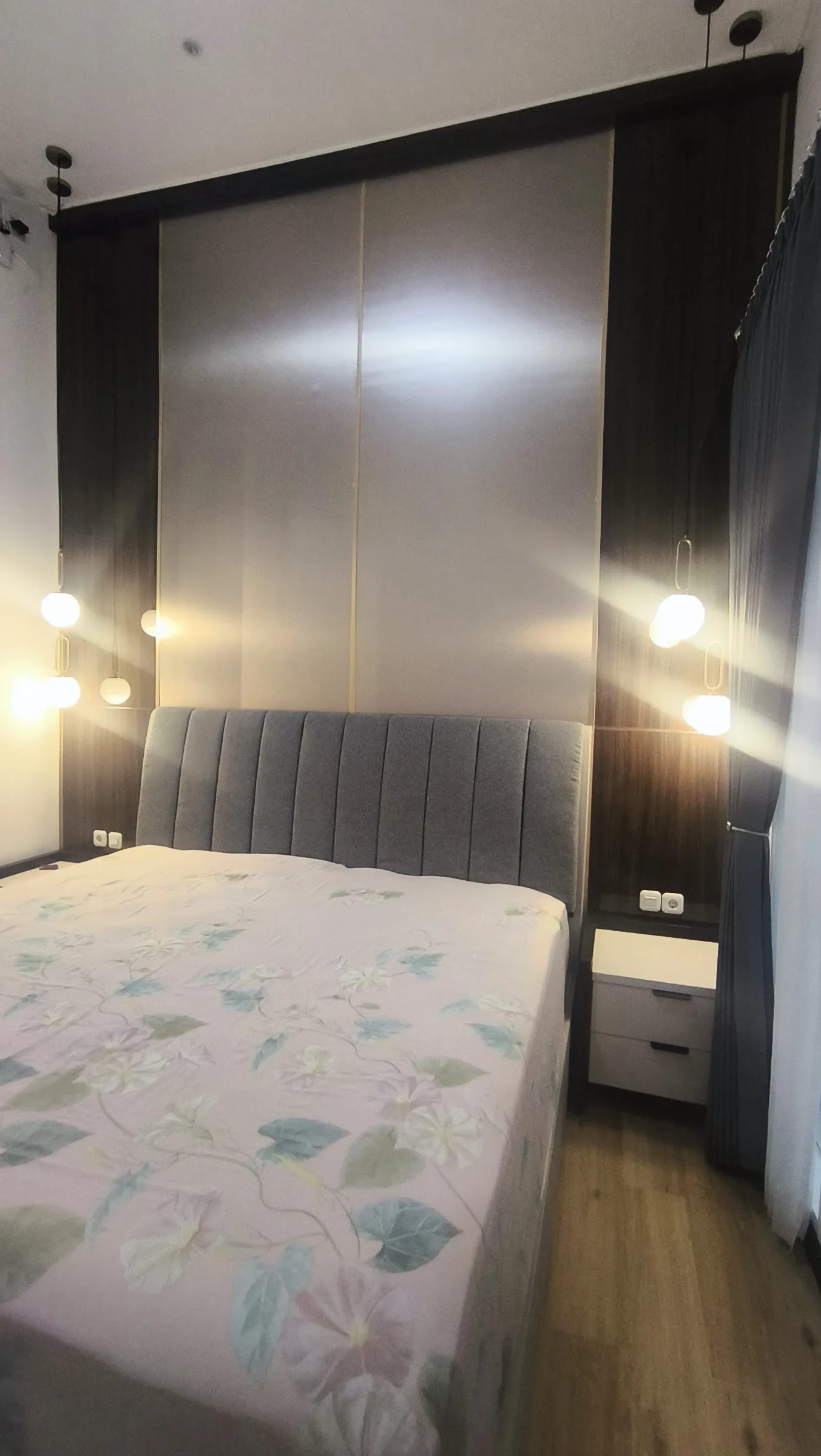 Kamar utama dengan tempat tidur, panel dinding abu-abu dan lampu dinding hitam